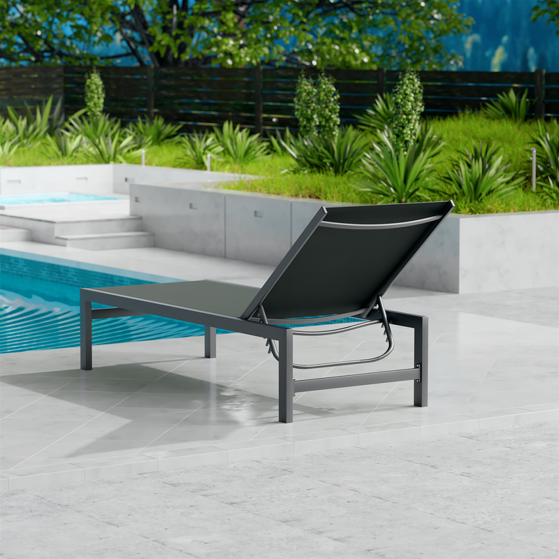A gray outdoor lounge chair with a dark frame is set on a sleek, light-colored tiled area beside a swimming pool. The background features lush greenery and a wooden fence, creating a serene outdoor relaxation spot. gray outdoor lounge chair, poolside lounge chair, outdoor relaxation furniture, dark-framed lounge chair.