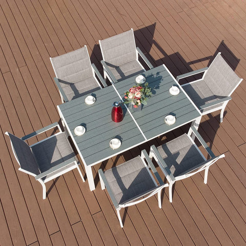 An overhead view of an outdoor dining setup featuring a rectangular composite - wood dining table with a light grey top and white frame. The table is surrounded by six chairs with grey fabric seats and backs, and is set with plates, cups, a red carafe, and a floral centerpiece on a wooden - deck patio. Rectangular Outdoor Dining Table with Chairs on Wooden Deck. rectangular dining table, composite wood table, outdoor dining set, white - framed table, wooden deck, grey - fabric chairs.