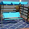 A blue-colored outdoor swing chair with a canopy is placed on a wooden deck by the water. The swing has a sturdy black metal frame and is positioned on a blue patterned rug. Next to the swing, there is a small black metal table with two glasses of orange beverages and a bunch of purple flowers. A potted plant adds a touch of greenery to the scene. Blue Swing Chair by the Water: A Serene Outdoor Retreat.
 blue swing chair, outdoor swing, swing with canopy, waterfront seating, patio furniture