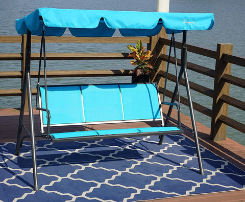 A blue-colored outdoor swing chair with a canopy is placed on a wooden deck by the water. The swing has a sturdy black metal frame and is positioned on a blue patterned rug. Next to the swing, there is a small black metal table with two glasses of orange beverages and a bunch of purple flowers. A potted plant adds a touch of greenery to the scene. Blue Swing Chair by the Water: A Serene Outdoor Retreat.
 blue swing chair, outdoor swing, swing with canopy, waterfront seating, patio furniture