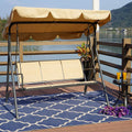 A beige - colored outdoor swing chair with a canopy is placed on a wooden deck by the water. The swing has a sturdy black metal frame and is positioned on a blue patterned rug. Next to the swing, there is a small black metal table with two glasses of orange beverages and a bunch of purple flowers. A potted plant adds a touch of greenery to the scene.Beige Swing Chair by the Water: A Serene Outdoor Retreat. beige swing chair, outdoor swing, swing with canopy, waterfront seating, patio furniture