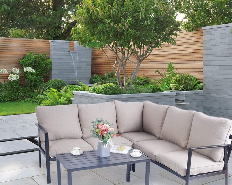 A cozy outdoor sectional sofa set with beige cushions and a black metal frame, placed on a stone - tiled patio. The set includes a coffee table adorned with a vase of flowers, cups, and a magazine. The background features a beautifully landscaped garden with greenery, a small tree, and a water feature, enhancing the serene outdoor ambiance.Serene Outdoor Sectional Sofa Set for Garden Retreats. outdoor sectional sofa, beige cushions, black metal frame, patio furniture, garden seating, outdoor coffee table
