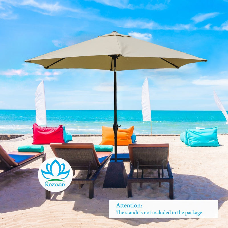A beige outdoor beach umbrella set on a sandy beach with a stunning blue - sea backdrop. Beneath the umbrella are lounge chairs, colorful bean bags, and blue cushions. A logo for "KOZYARD" is visible, and a notice states that the stand is not included in the package. KOZYARD Outdoor Beach Umbrella for Relaxing by the Sea. outdoor beach umbrella, KOZYARD, beach lounge, outdoor relaxation, umbrella stand not included