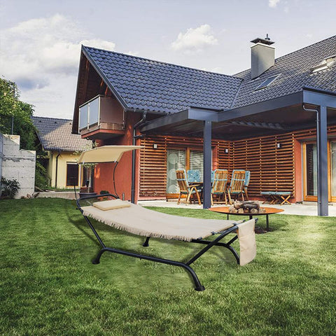 A beige outdoor hammock lounger with a black metal frame sits on a well - manicured green lawn. It features a small cushion, a sun canopy, and a side pocket. In the background, there is a modern house with wooden and orange - colored facades, outdoor furniture, and a fire pit area.Beige Outdoor Hammock Lounger for Garden Relaxation. outdoor hammock lounger, garden furniture, beige hammock, sun canopy, side pocket