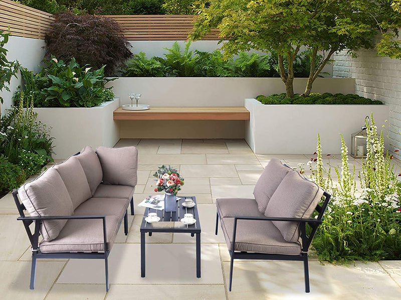 An outdoor seating arrangement featuring a set of modern sofas with beige cushions and black metal frames, paired with a black coffee table. The setup is placed on a tiled patio surrounded by lush greenery, with a wooden bench and a small tree in the background, creating a serene and inviting outdoor lounge space. Serene Outdoor Sofa Set for Relaxing Patio Spaces. outdoor sofa set, beige - cushioned sofas, black metal frame, patio furniture, outdoor lounge area
