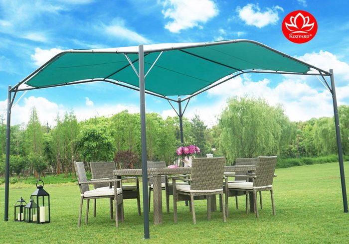 A KOZYARD Sun Shade Gazebo with a blue canopy is set on a lush green lawn. Beneath the gazebo, there is an outdoor wicker dining set with chairs and a table adorned with a vase of pink flowers. Lanterns are placed on the grass nearby, adding to the cozy outdoor ambiance. KOZYARD sun shade gazebo, blue canopy, outdoor wicker furniture, outdoor relaxation, garden lanterns.