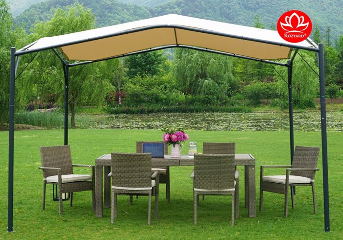 A KOZYARD sun shade gazebo with a beige canopy stands over an outdoor wicker dining set on a grassy area near a serene body of water surrounded by lush greenery. The gazebo provides a shaded spot for relaxation and dining.KOZYARD Sun Shade Gazebo for Outdoor Relaxation and Dining.KOZYARD sun shade gazebo, beige canopy, outdoor wicker furniture, shaded dining area, outdoor relaxation.