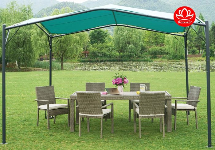 A KOZYARD sun shade gazebo with a blue canopy stands over an outdoor wicker dining set on a grassy area near a serene body of water surrounded by lush greenery. The gazebo provides a shaded spot for relaxation and dining. KOZYARD sun shade gazebo, blue canopy, outdoor wicker furniture, shaded dining area, outdoor relaxation.
