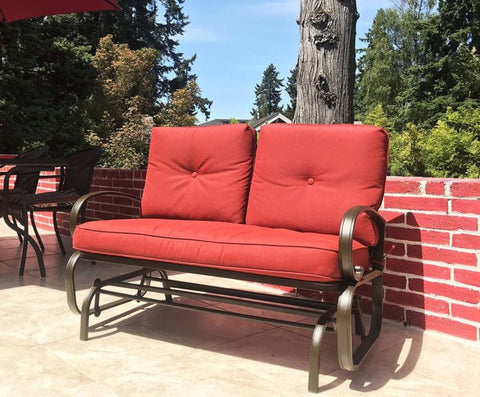 A vibrant red - cushioned glider chair placed on an outdoor patio. The chair features a sturdy metal frame with curved armrests, providing a comfortable spot for relaxation. The background includes greenery and a brick wall, enhancing the inviting outdoor atmosphere. Red - Cushioned Glider Chair for Outdoor Relaxation. red - cushioned glider chair, outdoor patio furniture, metal - framed chair, curved armrests, relaxation.