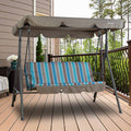 A Kozyard outdoor swing chair positioned on a wooden deck. The swing features a sturdy metal frame and a canopy in a neutral beige tone. The seat is adorned with a vibrant cushion showcasing a striped pattern in shades of blue, light blue, and brown. The background includes part of a house exterior, a railing, and some greenery, creating a cozy outdoor setting. Kozyard Outdoor Swing Chair: Relax in Style on Your Deck. Kozyard swing chair, outdoor patio swing, striped - cushion swing, swing with canopy, deck