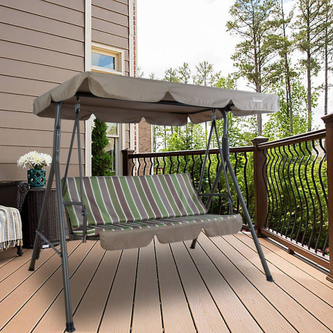 An outdoor patio swing chair from Kozyard is positioned on a wooden deck. The swing features a cushioned seat with green, brown, and white vertical stripes. Above it, a beige canopy provides shade, adding to the comfort. The background includes part of a house with a window, a decorative vase with white flowers, and a railing overlooking greenery, creating a relaxing outdoor setting. Kozyard Outdoor Swing Chair for Relaxing Patio Moments. Kozyard swing chair, outdoor patio swing, swing with canopy, striped 