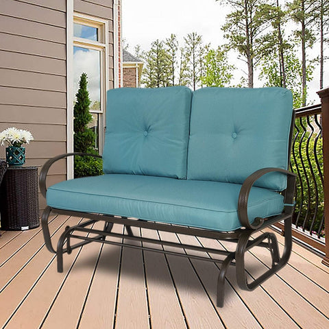 A glider chair with vibrant blue cushions is placed on a wooden deck, surrounded by a pleasant outdoor setting with greenery and a house facade in the background. The chair features a sturdy metal frame with curved armrests, providing a comfortable and stylish spot for relaxation.Blue - Cushioned Glider Chair for Outdoor Relaxation.blue - cushioned glider chair, outdoor furniture, metal frame, curved armrests, wooden deck
