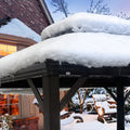 An outdoor gazebo with a dark - colored frame is blanketed in a thick layer of snow. Icicles hang from the edge of the roof, indicating a cold winter setting. In the background, there is a brick house with a lit window, and snow - covered trees and furniture, enhancing the wintry atmosphere. Outdoor Gazebo in Winter: Snow - Covered Serenity. outdoor gazebo, winter scene, snow - covered, icicles, winter atmosphere