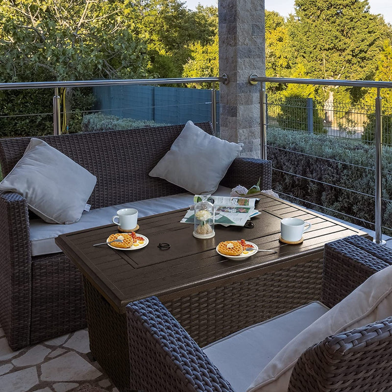 An outdoor patio scene featuring a dark - colored wicker fire pit table set. The table is adorned with cups, plates of waffles, a decorative jar, and a newspaper. Surrounding the table are wicker sofas and chairs with light - colored cushions, set against a backdrop of greenery and a railing, creating a cozy and inviting outdoor relaxation area. Cozy Outdoor Wicker Fire Pit Table Set for Relaxation. outdoor fire pit table, wicker patio set, cozy outdoor seating, fire pit furniture, patio relaxation area.