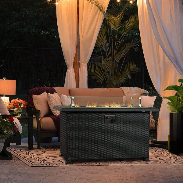 An outdoor wicker - styled fire pit table with a glass - enclosed flame area, situated in a cozy patio setting. The table is placed on an ornate rug, surrounded by a comfortable beige sofa adorned with pillows and throws. Softly lit curtains and greenery add to the inviting atmosphere. Cozy Outdoor Wicker Fire Pit Table for Patio Ambiance. outdoor fire pit table, wicker fire pit, patio fire table, glass - enclosed flame, outdoor living space