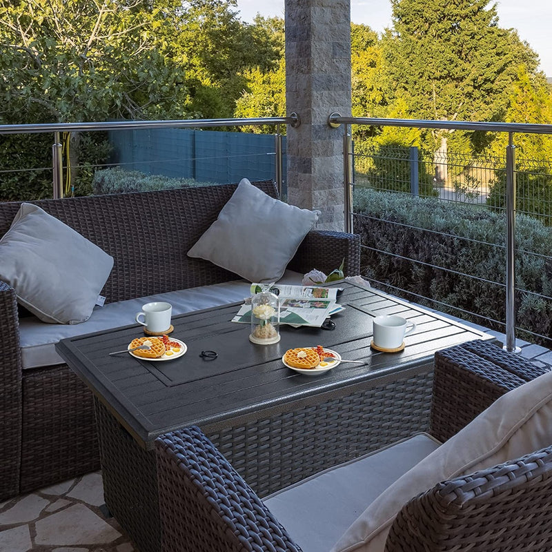 An outdoor patio scene featuring a dark - colored wicker fire pit table set. The table is adorned with cups, plates of waffles, a decorative jar, and a newspaper. Surrounding the table are wicker sofas and chairs with light - colored cushions, set against a backdrop of greenery and a railing, creating a cozy and inviting outdoor relaxation area. Cozy Outdoor Wicker Fire Pit Table Set for Relaxation. outdoor fire pit table, wicker patio set, cozy outdoor seating, fire pit furniture, patio relaxation area.