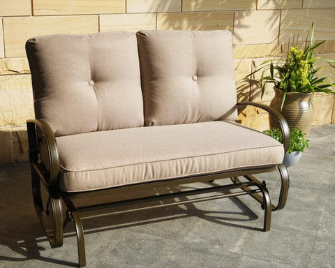 A glider chair with beige cushions is positioned on an outdoor stone - tiled area, next to a potted plant and a stone wall. The chair has a sturdy metal frame with curved armrests, providing a comfortable and stylish spot for relaxation.Beige - Cushioned Glider Chair for Outdoor Relaxation.beige - cushioned glider chair, outdoor furniture, metal - framed chair, curved armrests, relaxation
