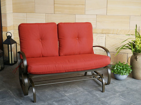 A glider chair with vibrant red cushions is placed on a stone - tiled floor next to decorative plants and a lantern. The chair features a sturdy metal frame with curved armrests, providing a comfortable and stylish spot for relaxation. Red - Cushioned Glider Chair for Relaxing Moments.red - cushioned glider chair, metal - framed glider, outdoor relaxation, curved armrests.